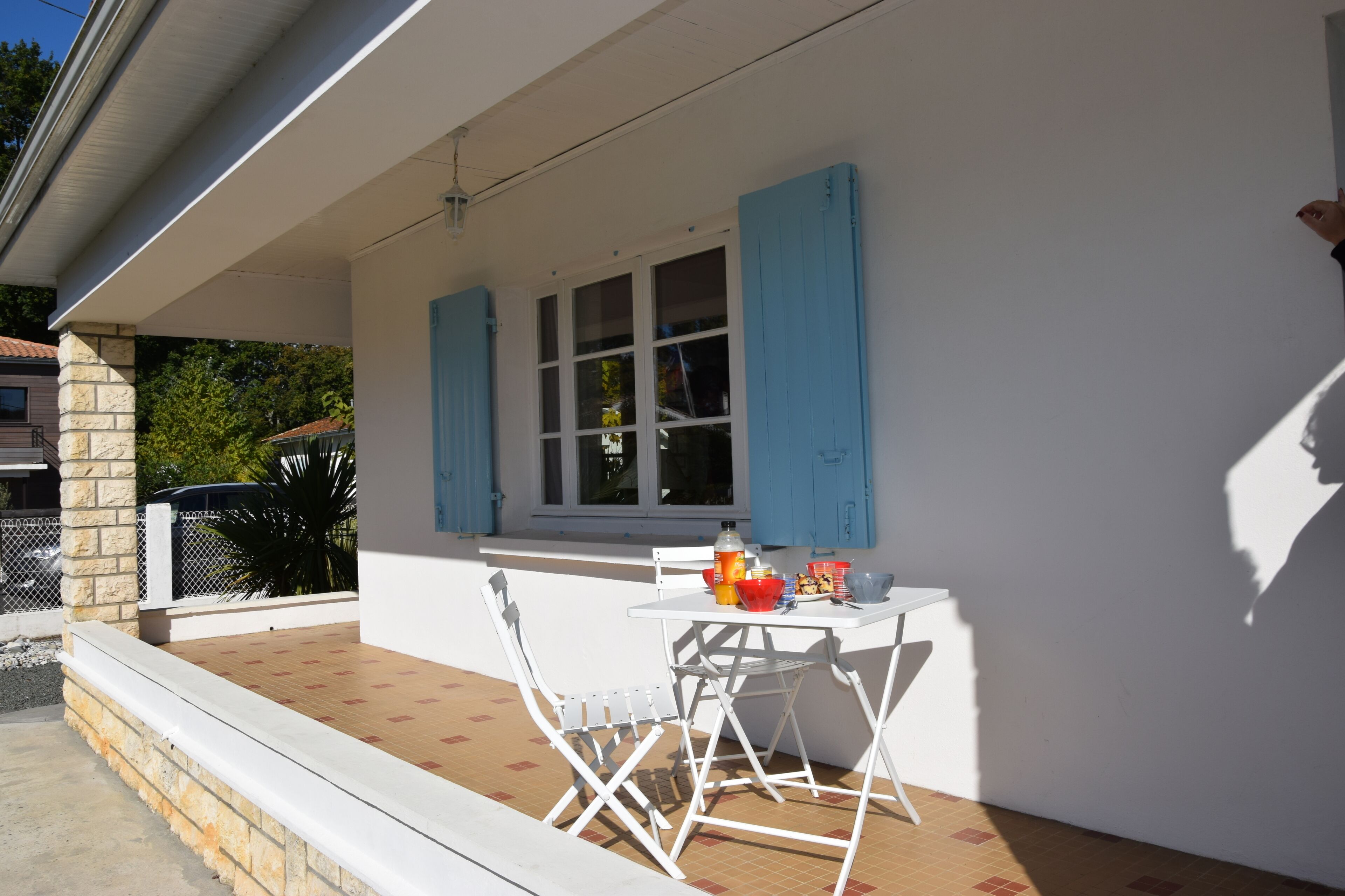 Une terrasse couverte avec une table et des chaises blanches, une fenêtre avec des volets bleus et un pilier en pierre.