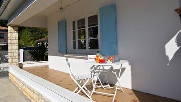 Une terrasse couverte avec une table et des chaises blanches, une fenêtre avec des volets bleus et un pilier en pierre.