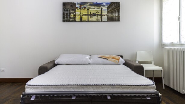 A neatly made bed with a mattress and pillows, a white chair, and a radiator.
