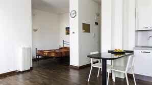 A modern hotel room with a wooden floor, a bed with a patterned blanket, a small table with chairs, a clock on the wall, and a kitchen area with a fruit bowl.