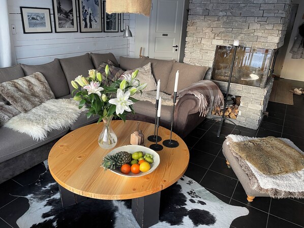 A living room with a sofa, a round wooden table with fruit, a vase with flowers, and a stone fireplace.