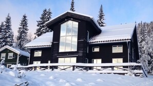 A large, dark wooden cabin with a prominent central window and a sloped roof, surrounded by snow-covered trees.