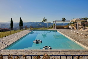A rectangular swimming pool with two inflatable rings, surrounded by a stone deck, a fenced area with lounge chairs, and a view of mountains in the background.