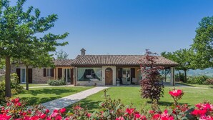 A stone house with a tiled roof, a covered porch, and a lawn with pink flowers. - Casa Emanuela - Private villa, 4 bedrooms, pool, A/C, wifi, pets, Marche (Fermignano)