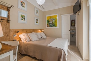 A bedroom with a wooden headboard, a bed with a beige quilt, a bedside table with a lamp, a TV mounted on the wall, and a bathroom visible through an open door.