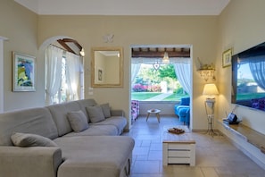 A living room with a sofa, a small table, a TV, and a view of a garden through an open door. - Casa Emanuela - Private villa, 4 bedrooms, pool, A/C, wifi, pets, Marche (Fermignano)