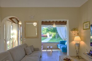 Ein Wohnzimmer mit einer Couch, einem kleinen Tisch, einer Lampe, einem Spiegel und Blick auf einen Garten durch eine offene Tür.