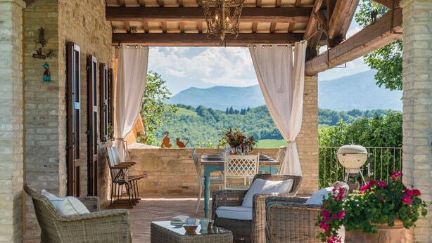 A covered outdoor seating area with wicker furniture, a table with a vase, and a view of green hills. - Casa Emanuela - Private villa, 4 bedrooms, pool, A/C, wifi, pets, Marche (Fermignano)