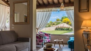 A living room with a grey sofa, a floral armchair, a small round table with books and a bowl of fruit, and a view of a garden through a window with sheer curtains. - Casa Emanuela - Private villa, 4 bedrooms, pool, A/C, wifi, pets, Marche (Fermignano)