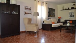A living room with a dark wooden cabinet, beige armchair, and a sofa with orange cushions. There are framed pictures on the wall and a lamp on a side table. - Villa with private pool in Pinar de los Franceses (WIFI) (Chiclana De La Frontera)