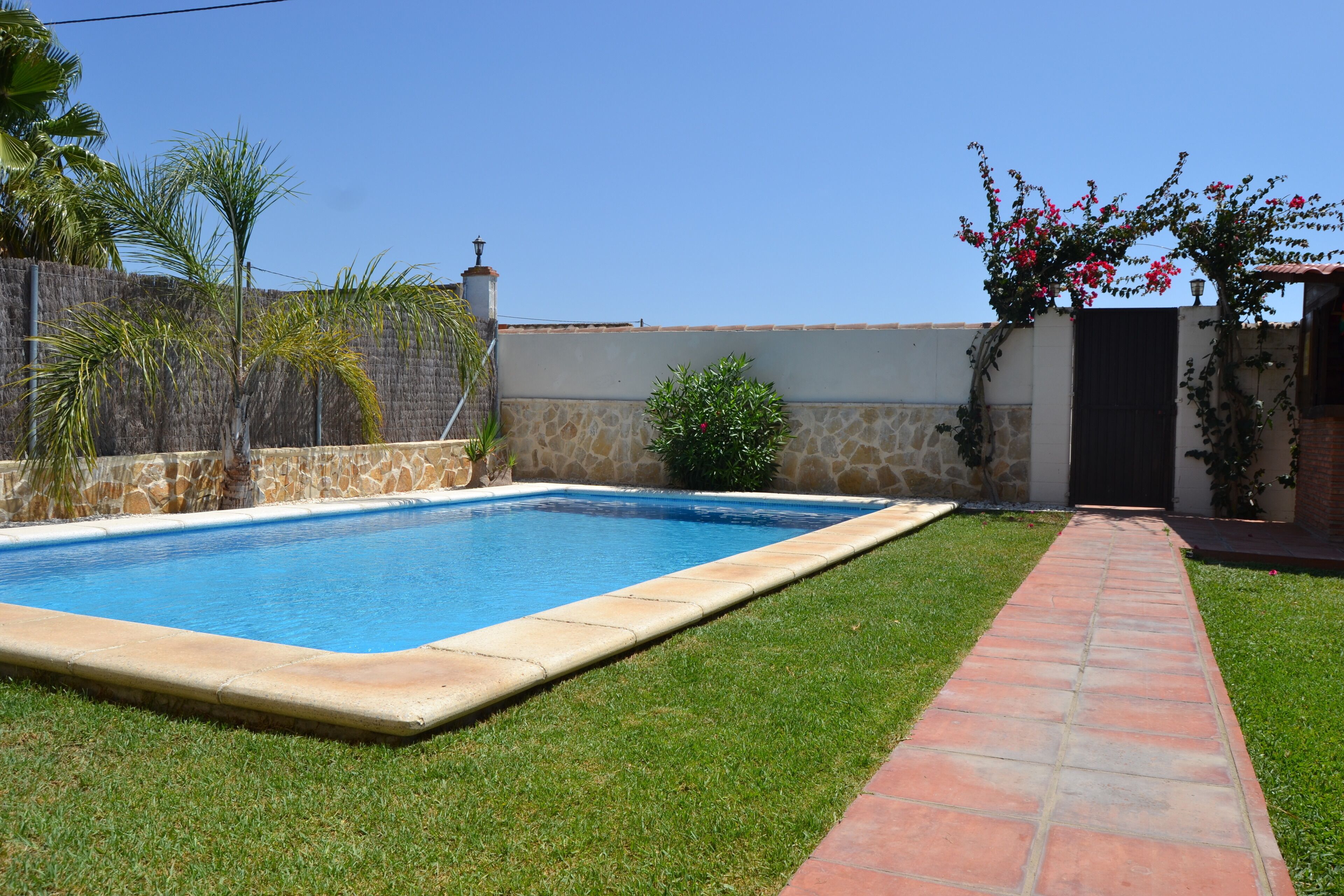 Une piscine rectangulaire bordée de pierres, entourée d’une pelouse bien entretenue et d’un chemin en briques menant à un bâtiment.