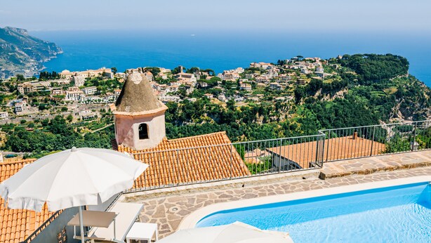 A pool area with white umbrellas and lounge chairs, overlooking a coastal village with terracotta-roofed buildings and a tower. - Apt Emma in Villa Laura near Ravello (scala)