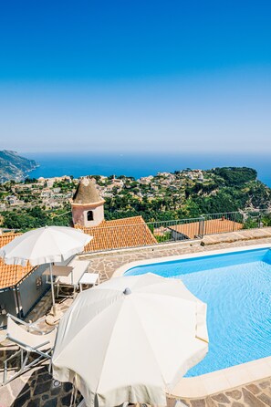 A pool area with white umbrellas and lounge chairs, overlooking a coastal village with terracotta-roofed buildings and a tower. - Apt Emma in Villa Laura near Ravello (scala)