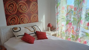 A neatly made bed with a white bedspread and red pillows, a decorative wall tapestry, a small bedside table with a lamp, and a floral curtain. - Apartment/ flat - LUMIO (Lumio)
