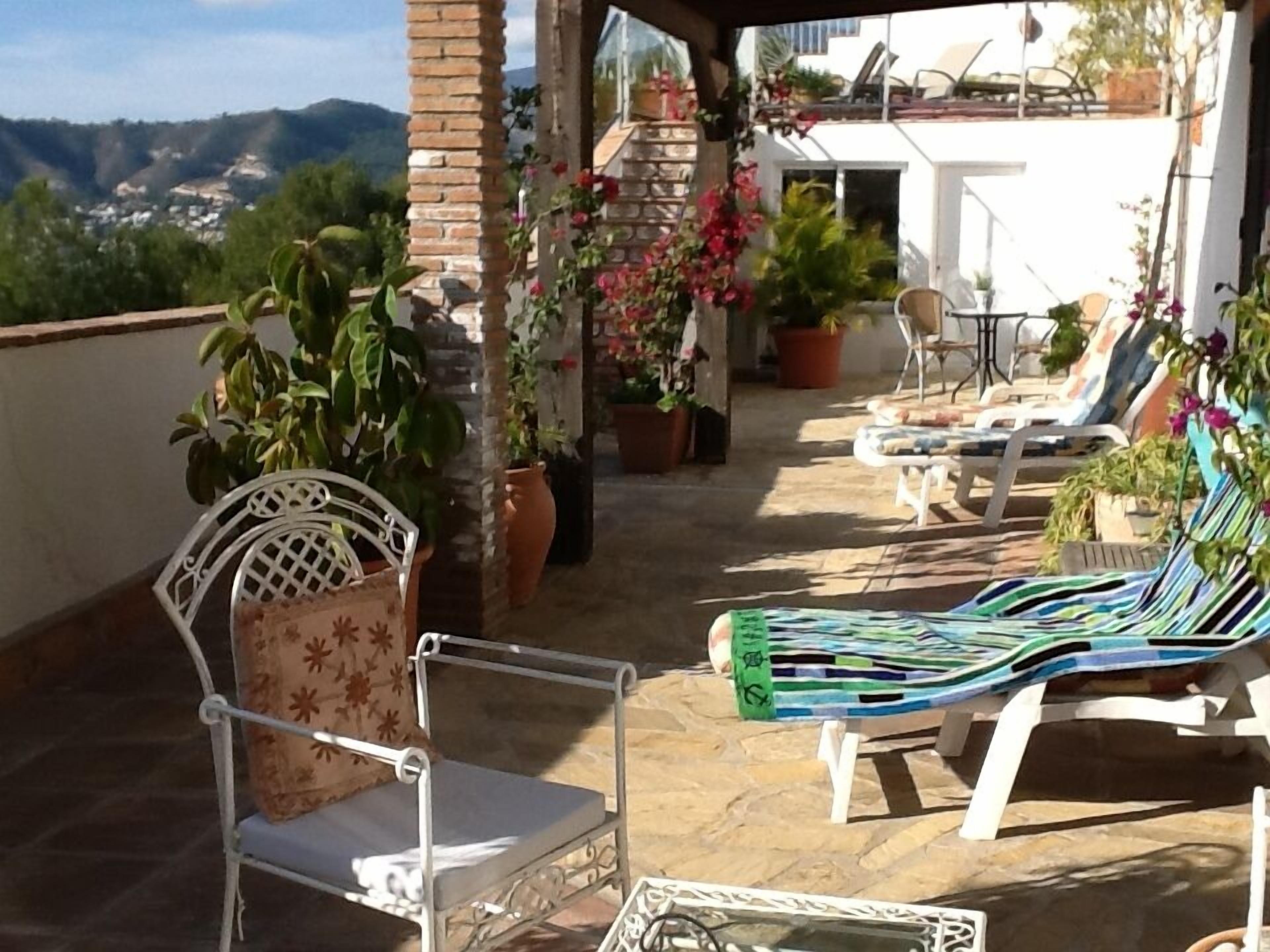 Un espacio de estar al aire libre cubierto con sillas y una mesa, rodeado de plantas y flores en macetas.