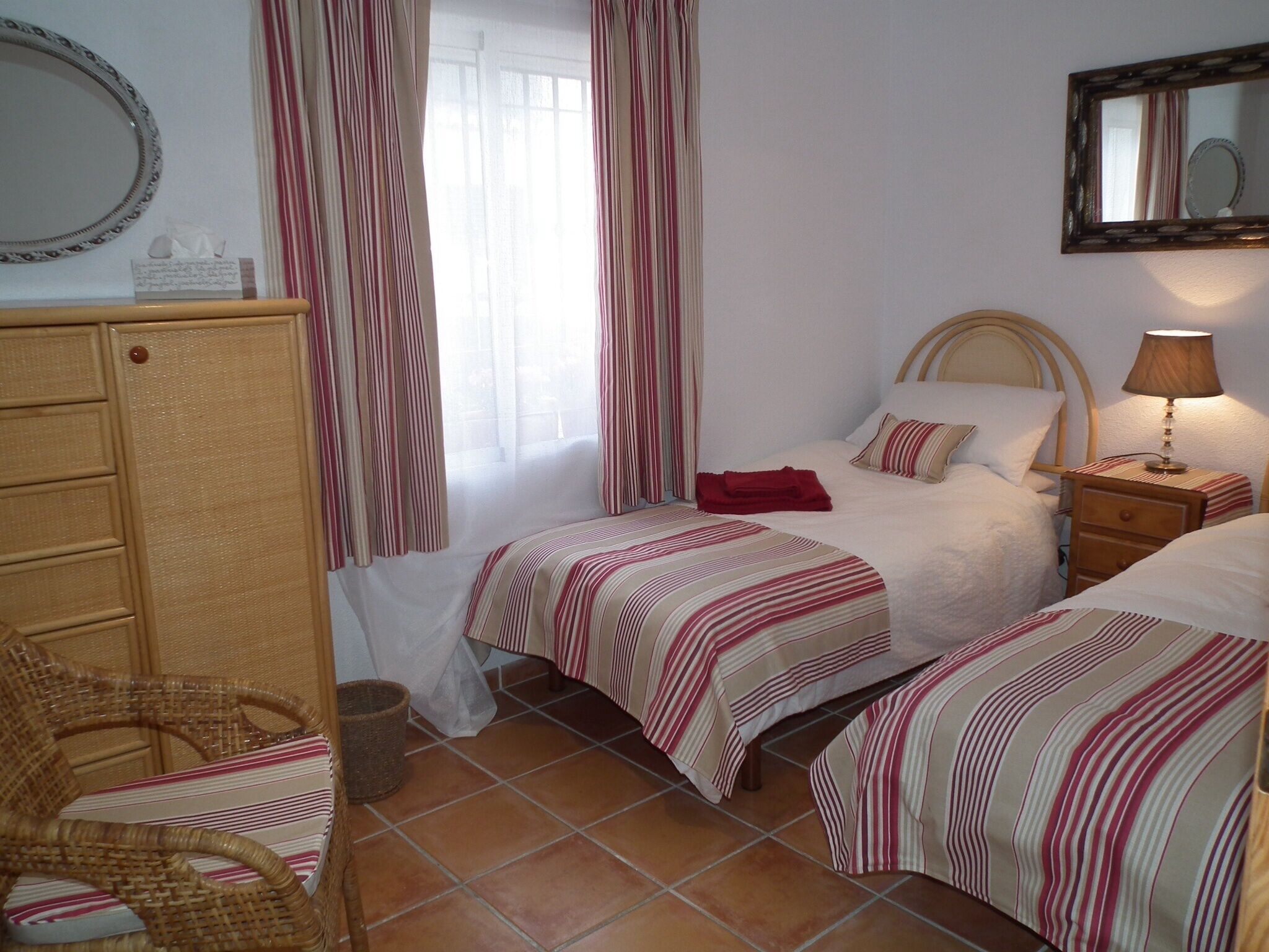 A bedroom with two beds, a wooden dresser, a wicker chair, a bedside table with a lamp, and a mirror on the wall.