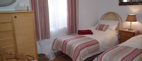 A bedroom with two beds, a wooden dresser, a wicker chair, a bedside table with a lamp, and a mirror on the wall.
