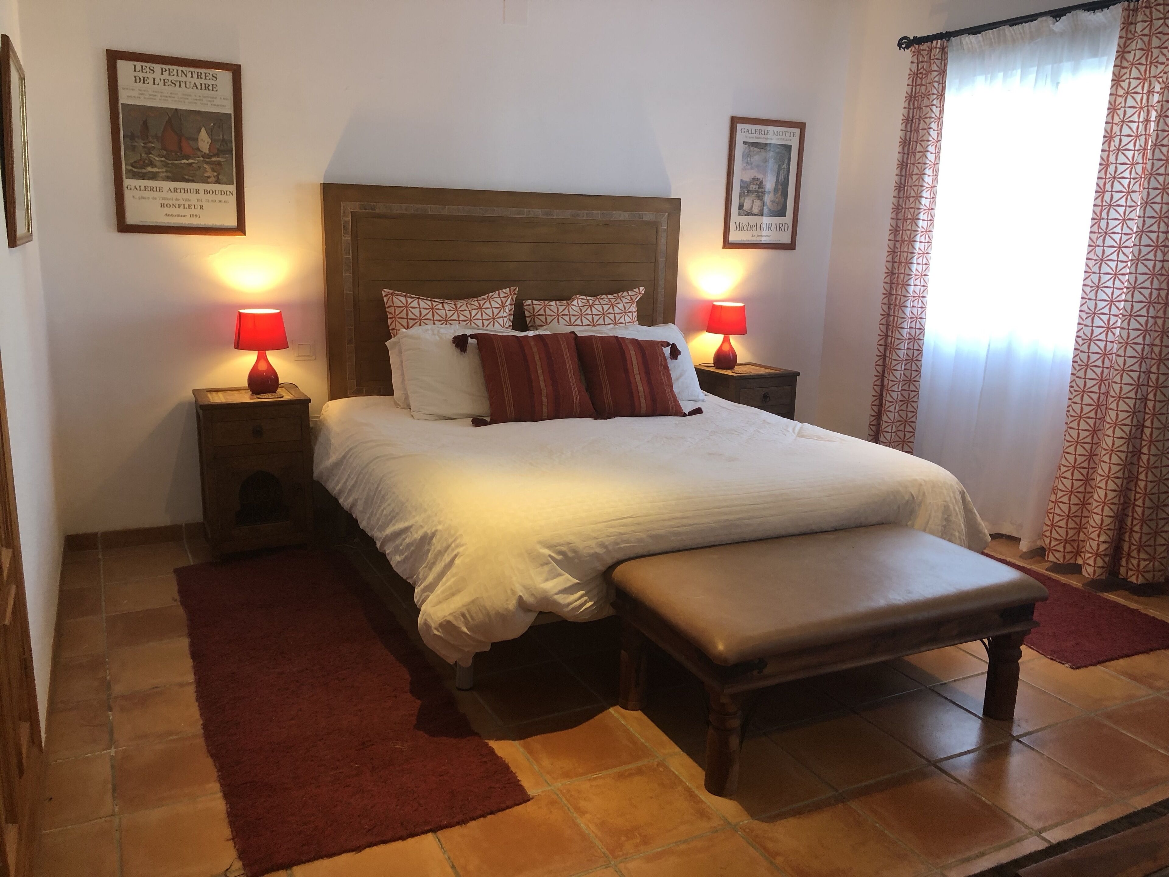 Un lit bien fait, avec une tête de lit en bois, deux tables de chevet avec des lampes, un tapis rouge et deux tableaux encadrés accrochés aux murs.