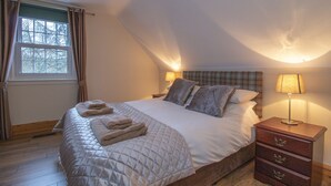 A bedroom with a bed, pillows, a bedside table, a lamp, a window with curtains, and a wooden floor.
