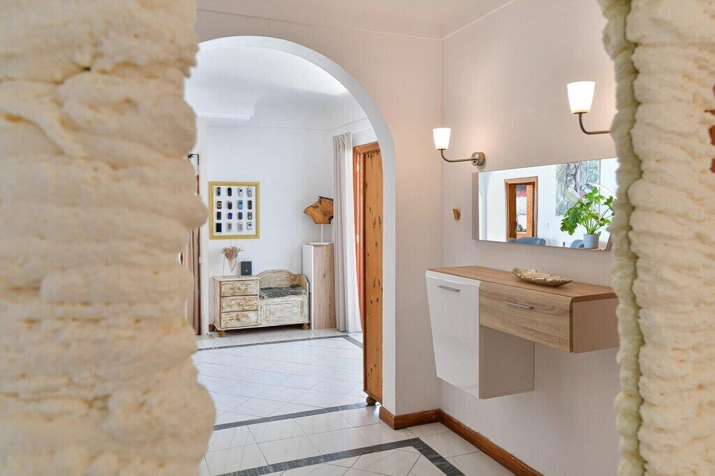 Un couloir avec un sol carrelé de carreaux blancs, une porte en bois, une commode blanche et un miroir agrémenté d’appliques murales.