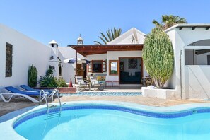 A poolside area with a clear blue pool, lounge chairs, and a building with a white exterior and a tiled roof.