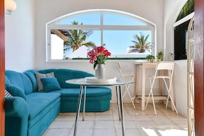 Een hoekappartement met een blauwgroene bank, een klein rond tafeltje met een bloevas, een witte stoel, en uitzicht op palmbomen door een groot rondboogvenster.