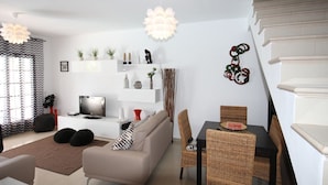 Un salon moderne avec un canapé, une table à manger et des chaises. On y trouve un téléviseur posé sur une console blanche, ainsi que des objets décoratifs sur des étagères.