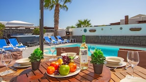 Un espace repas aménagé au bord de la piscine, avec une table dressée pour deux personnes, comprenant une assiette de fruits, une bouteille de vin et une vue sur la piscine et les chaises longues.