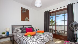 Une chambre à coucher avec un lit, une table de chevet, une plante en pot, un ventilateur et une fenêtre avec des rideaux.