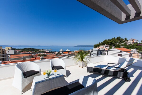 Une terrasse sur le toit, meublée de mobilier en osier blanc, avec une table sur laquelle se trouve un verre de jus d’orange, et une vue sur la mer et les bâtiments.