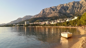 A coastal scene with a boat on the shore, a mountain in the background, and buildings along the coastline. - Luxury villa Palladium in the center with * heated pool, * penthouse and * whirlp. (Makarska)