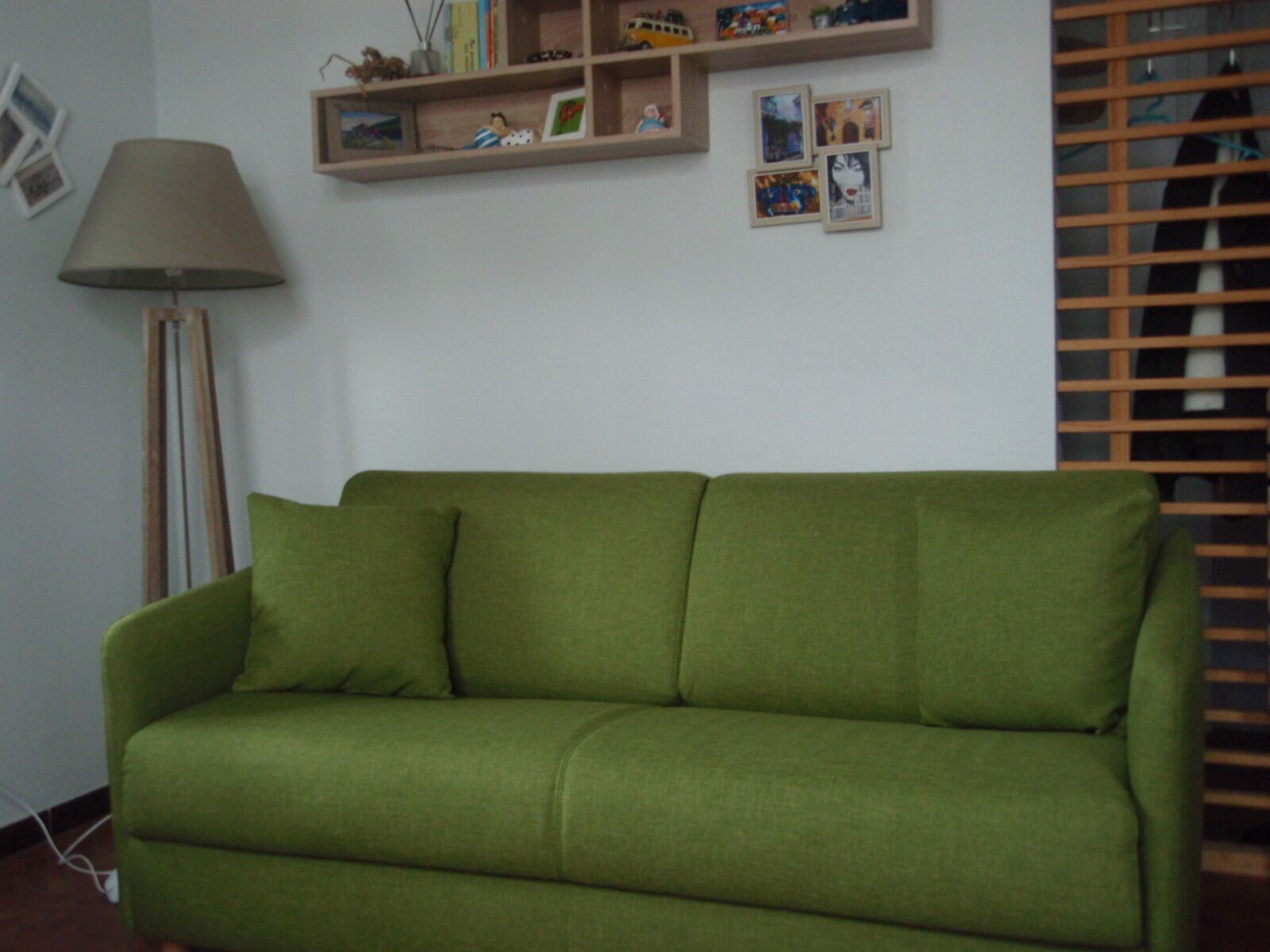 Un divano verde in una stanza con una lampada, mensole con quadri e piccoli oggetti, e una finestra con persiane.