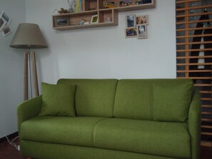 Ein grünes Sofa in einem Raum mit einer Lampe, Regalen mit Bildern und Kleinigkeiten sowie einem Fenster mit Jalousien.