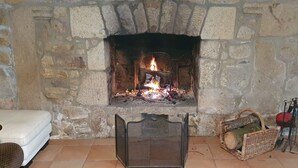 A stone fireplace with a fire, a wicker basket, and a metal screen in a room with a white sofa. - 17th-century farmhouse with large wooded park - quiet and charming, walking distance to town center and port (Étel)