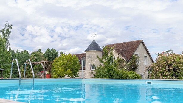 Una piscina con una escalera metálica, rodeada de árboles y un edificio con torre.
