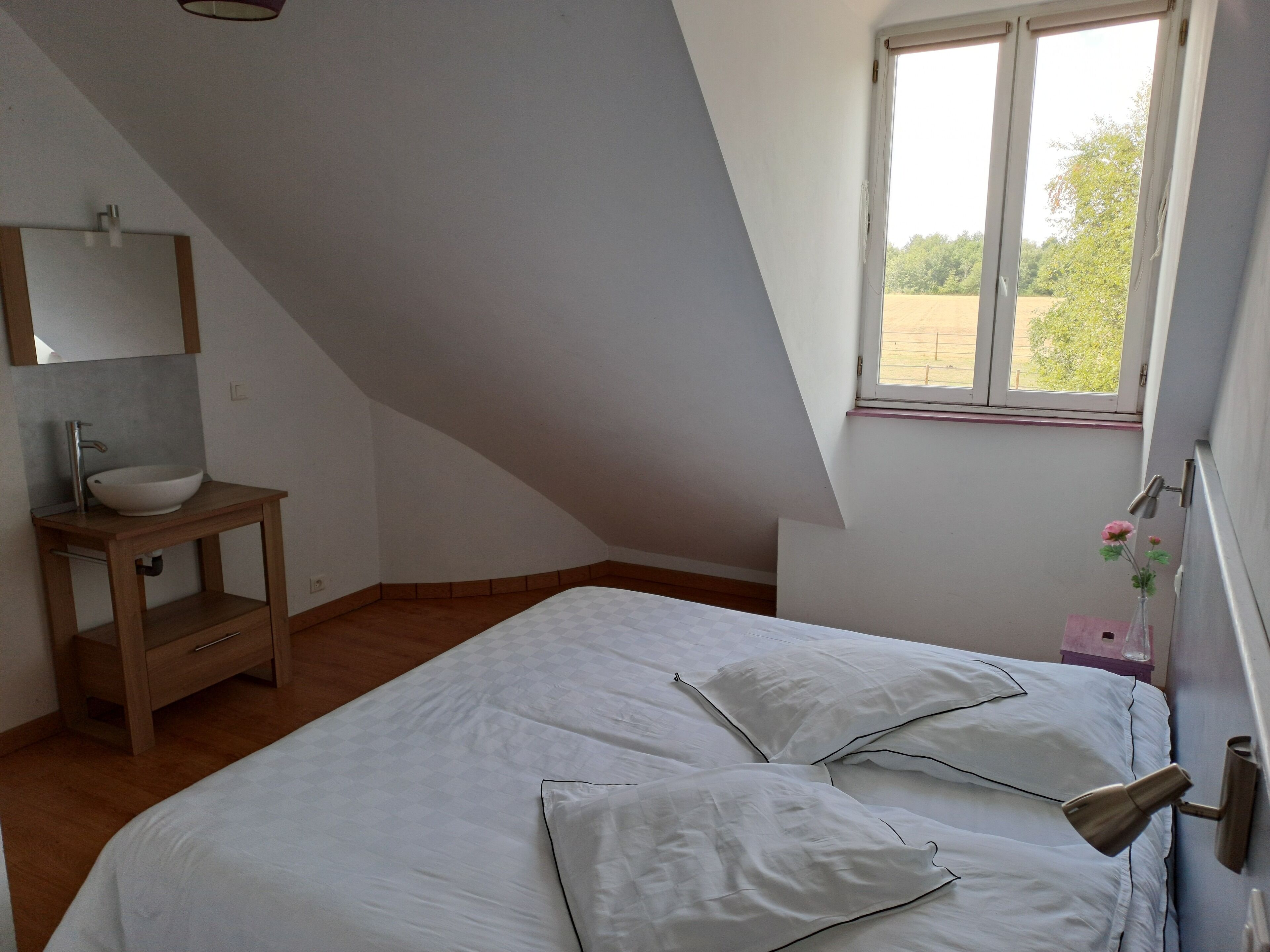 Ein Schlafzimmer mit schrägverdeckter Decke, einem Nachttisch aus Holz mit weißem Waschbecken, einem Bett mit weißer Bettwäsche und einem Fenster mit Blick auf Grünflächen.