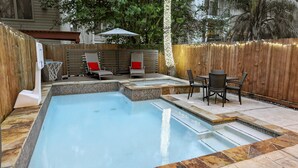 Piscina con borde de piedra y zona de cubierta de madera con sillas y una mesa.