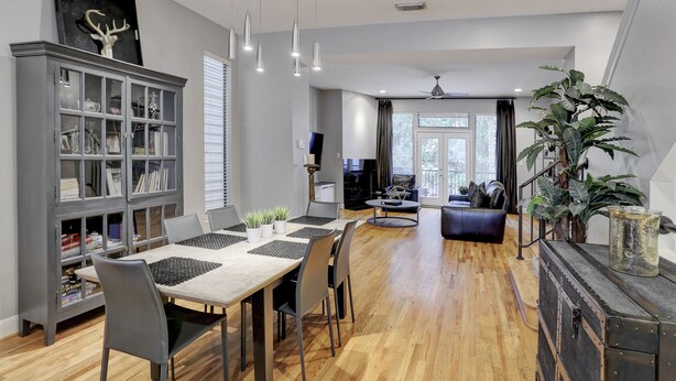 Una sala de estar moderna con comedor, suelo de madera, muebles grises y una planta grande.