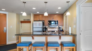 A kitchen with wooden cabinets, a stainless steel refrigerator, a microwave, and a sink. There are three blue bar stools and two pendant lights above the counter. - Front Corner at Base: 3 King Beds, Deck, Views (Winter Park)