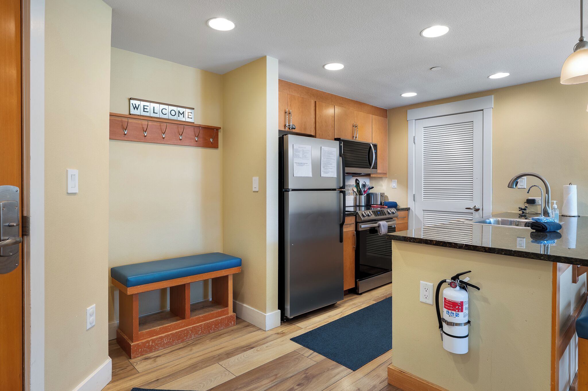 A compact kitchen with wooden cabinets, a stainless steel refrigerator, a microwave, and a built-in oven. There is a bench with a blue cushion, a welcome sign, and a fire extinguisher mounted on the wall.