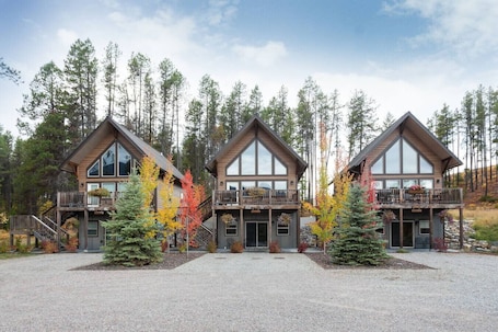 A two-story log cabin with a balcony, surrounded by a gravel driveway and autumn-colored trees.