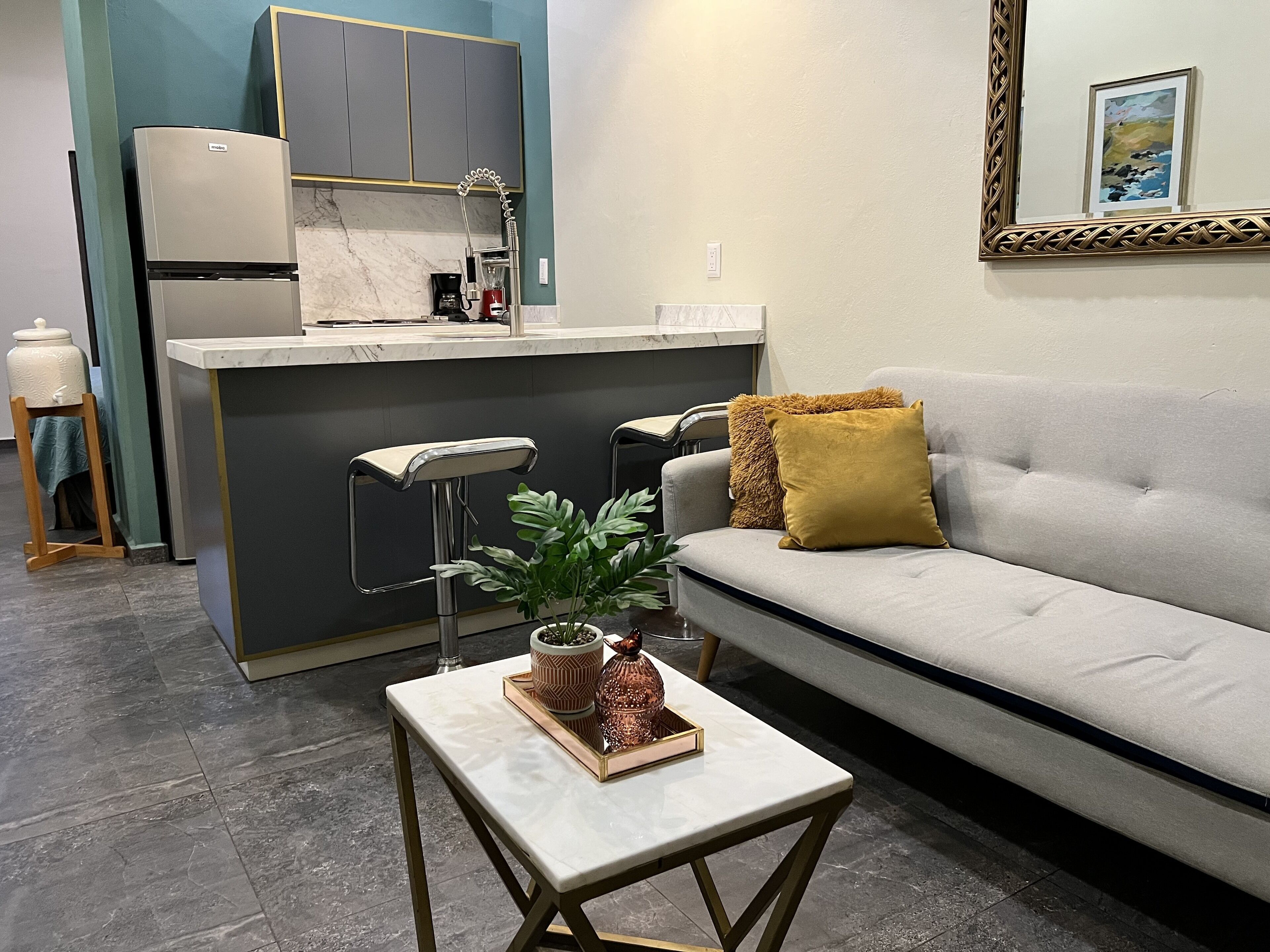 A modern kitchen with a built-in island, bar stools, and a grey sofa with a mustard throw pillow.