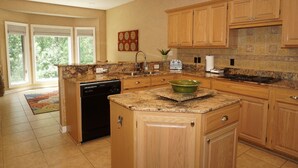 A kitchen with wooden cabinets, granite countertops, a dishwasher, and a large window with a view of greenery.