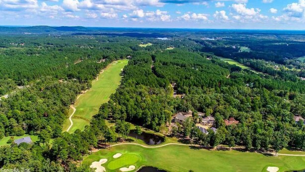 An aerial view of a golf course surrounded by dense forests, with a clear sky and distant hills.