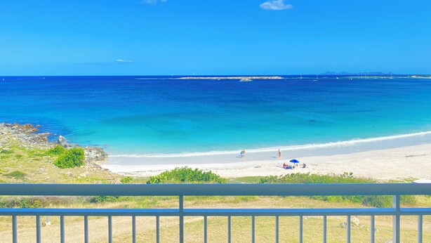 A beach view from a balcony with a railing, clear blue sky, and calm sea.