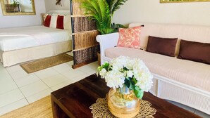 A room with a bed, a sofa, a coffee table with a vase of flowers, a woven basket, and a painting on the wall.
