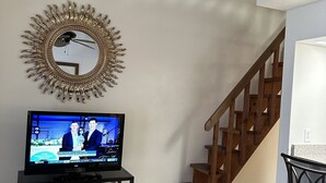 Ein Wohnzimmer mit einem Fernseher auf einem schwarzen Standfuß, einem Spiegel mit Sonnenstrahlendesign, einer Holztreppe und einem schwarzen Sessel.