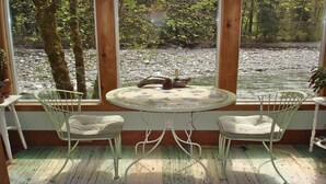 Une pièce dotée d’une grande fenêtre donnant sur un ruisseau aux rochers apparents, de deux chaises blanches et d’une table ronde recouverte d’une nappe à motifs floraux.