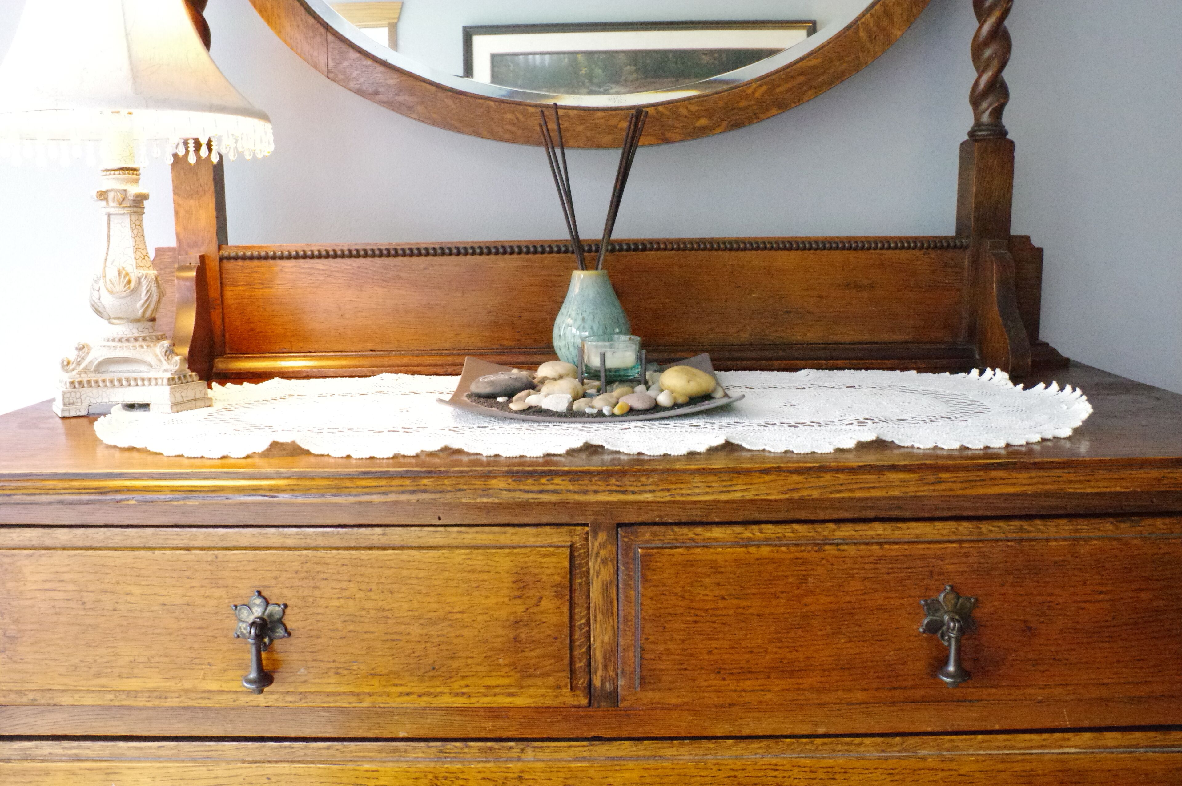 A wooden dresser with a mirror above it, a lace doily, a vase with stones, and a lamp.
