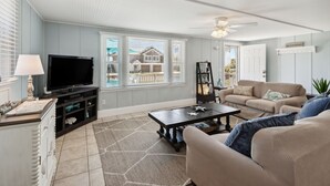 A living room with a television, sofa, coffee table, and a view of a house through large windows.